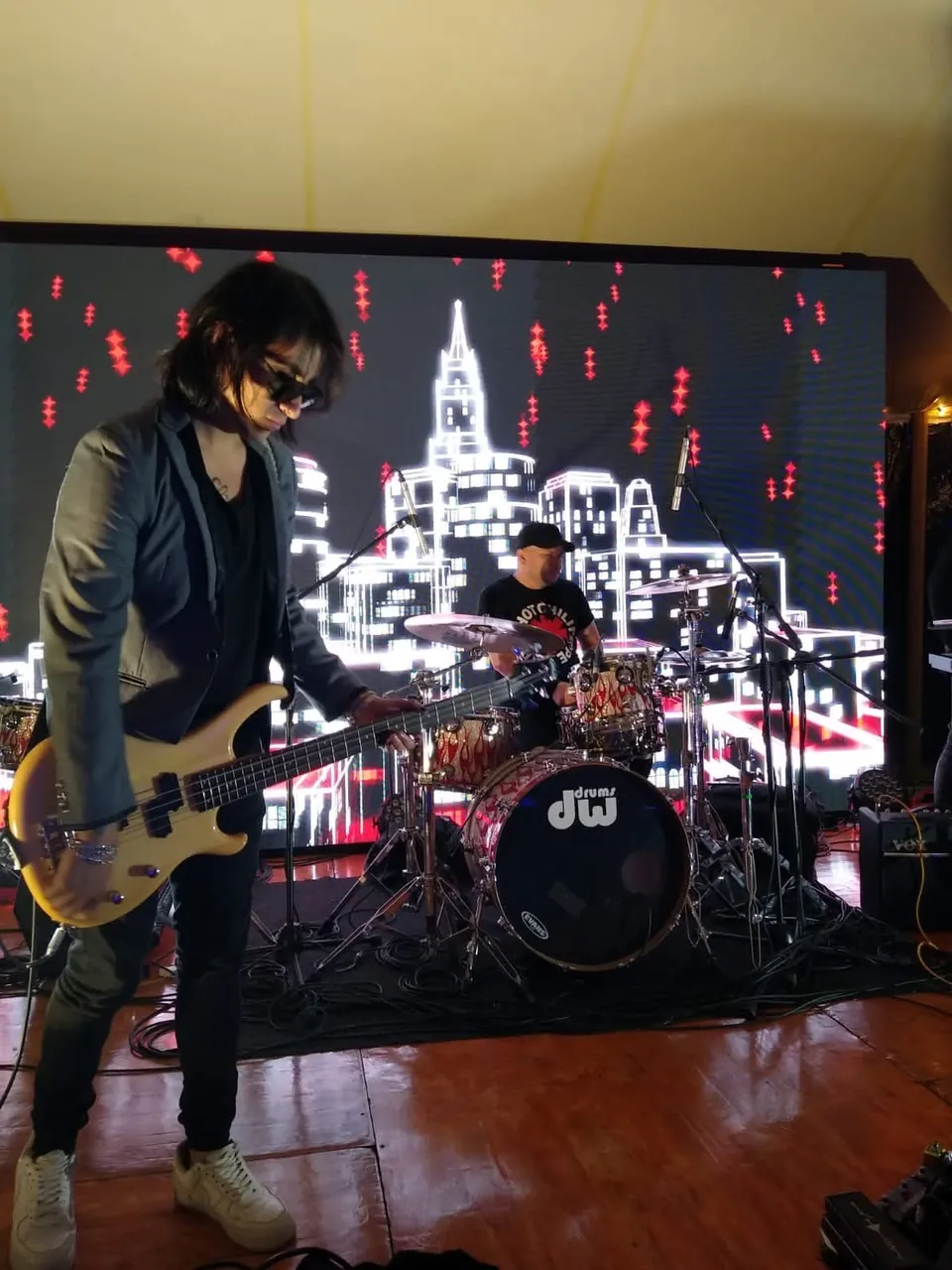 Bajista de la banda toca en vivo sobre la tarima frente a una pantalla LED con una ciudad nocturna.