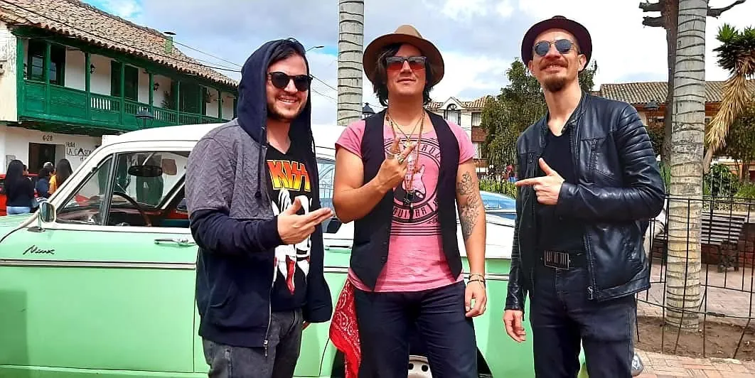 Mateo Acevedo junto a integrantes de su banda posando frente a un auto clásico en una plaza colonial.