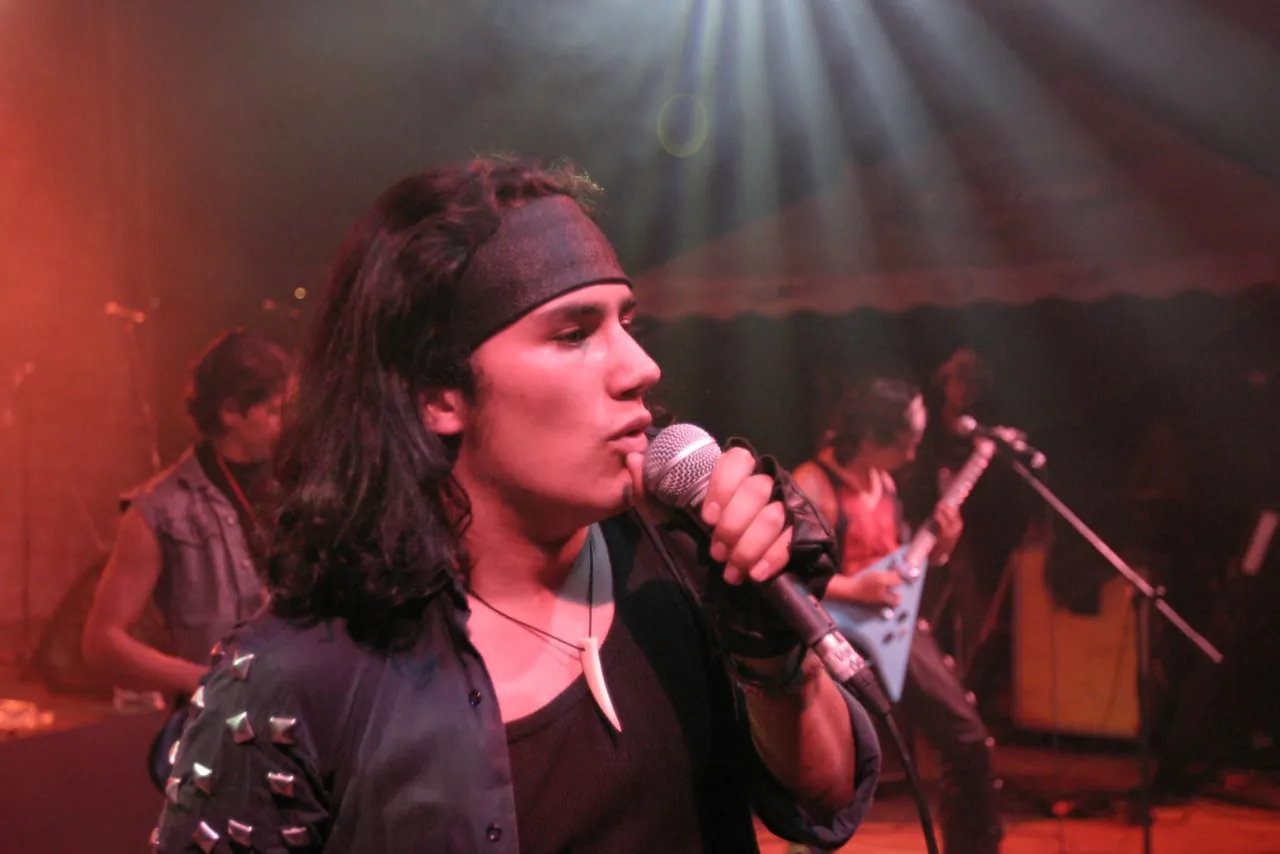 Primer plano de Mateo Acevedo cantando con chaqueta tachonada y su banda al fondo en un concierto de rock.