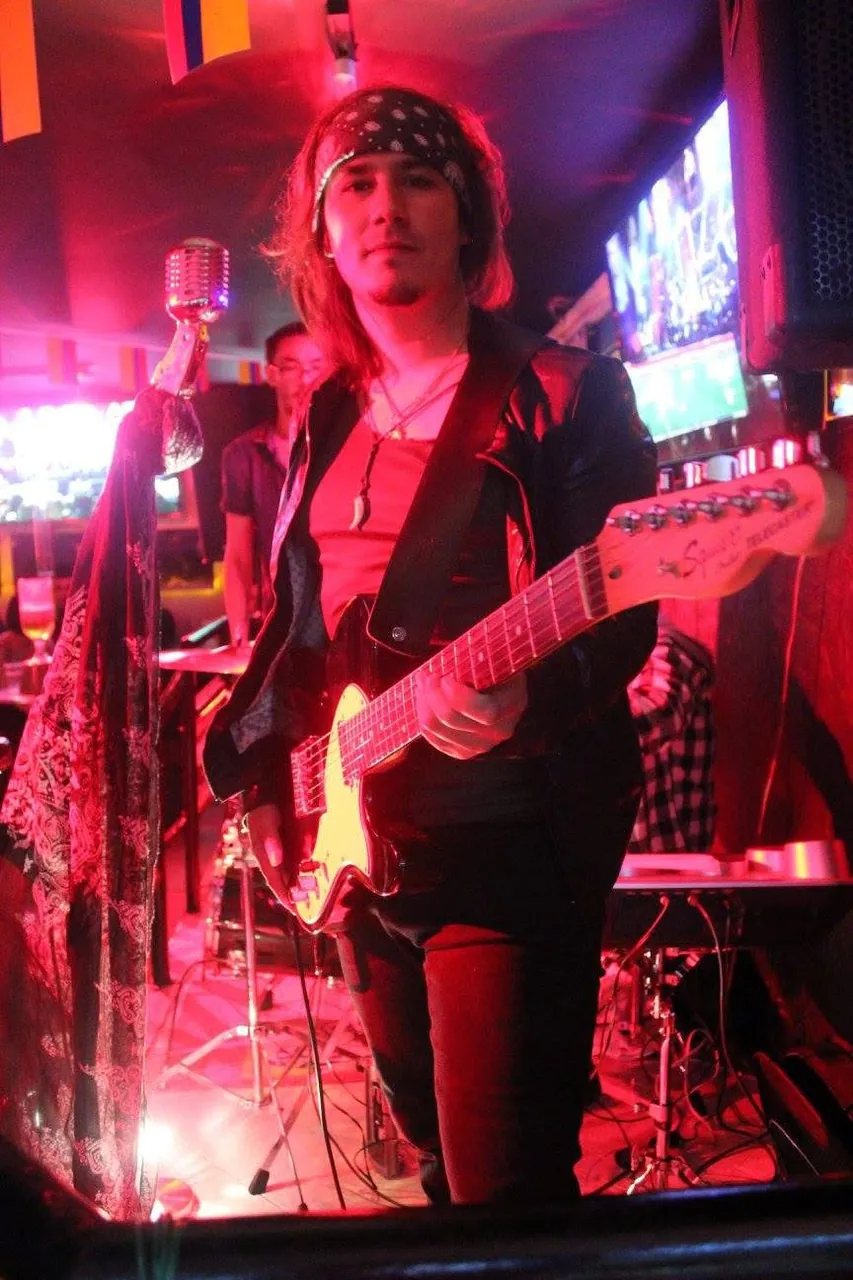 Retrato de Mateo Acevedo con bandana y guitarra junto al micrófono vintage bajo luces rojas en vivo.