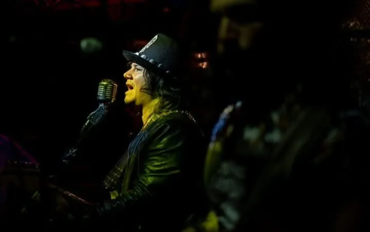 Retrato de Mateo Acevedo cantando frente a un micrófono vintage bajo un foco durante un show en vivo.