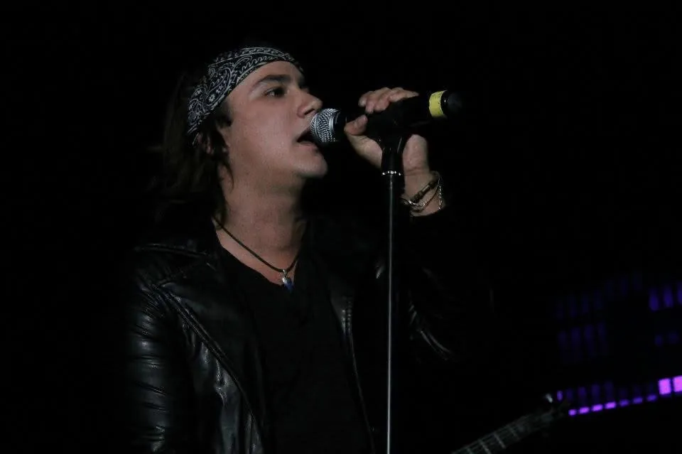 Mateo Acevedo cantando con micrófono y pañoleta, vestido de cuero negro, en una presentación de rock en vivo.