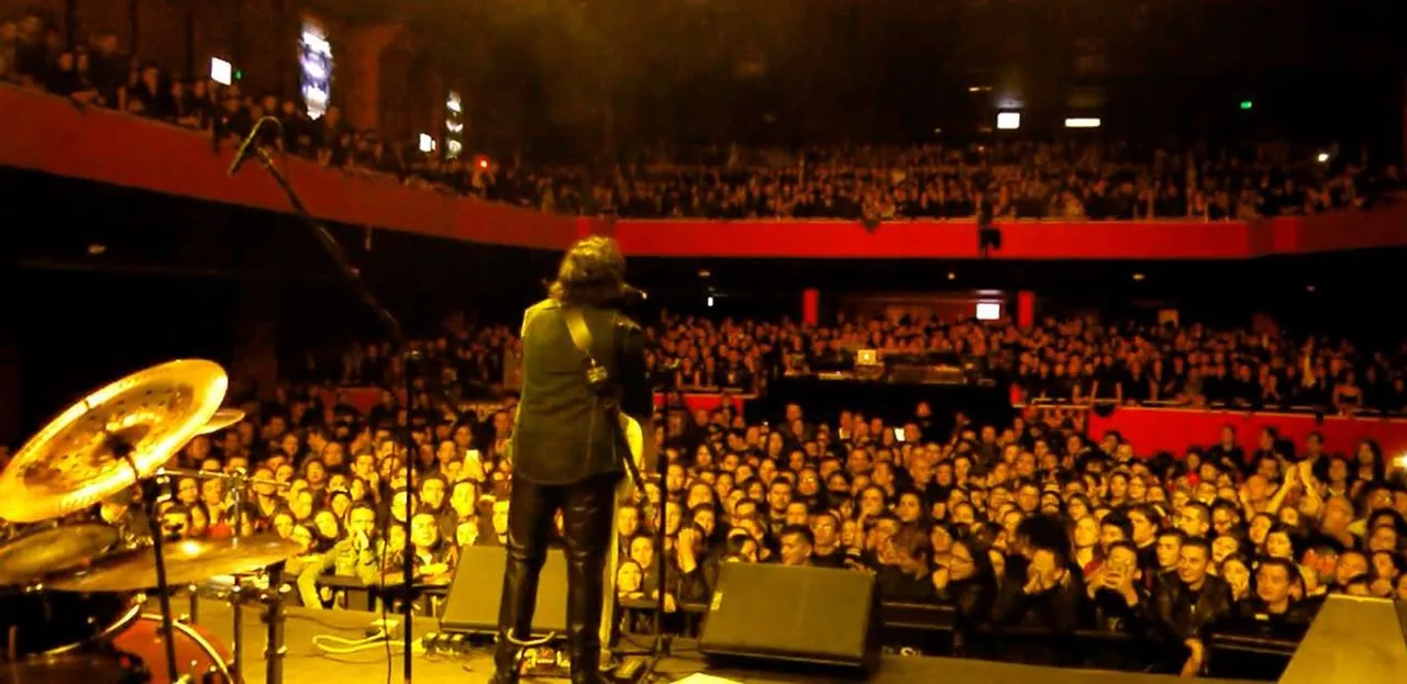 Mateo Acevedo de espaldas frente a un público multitudinario durante un concierto en un gran teatro.