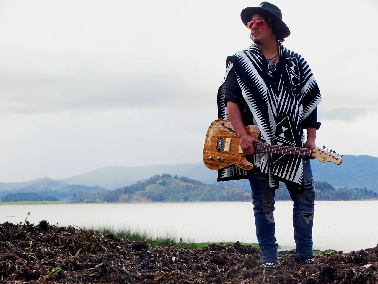 Mateo Acevedo con poncho y guitarra eléctrica frente a una laguna en Colombia