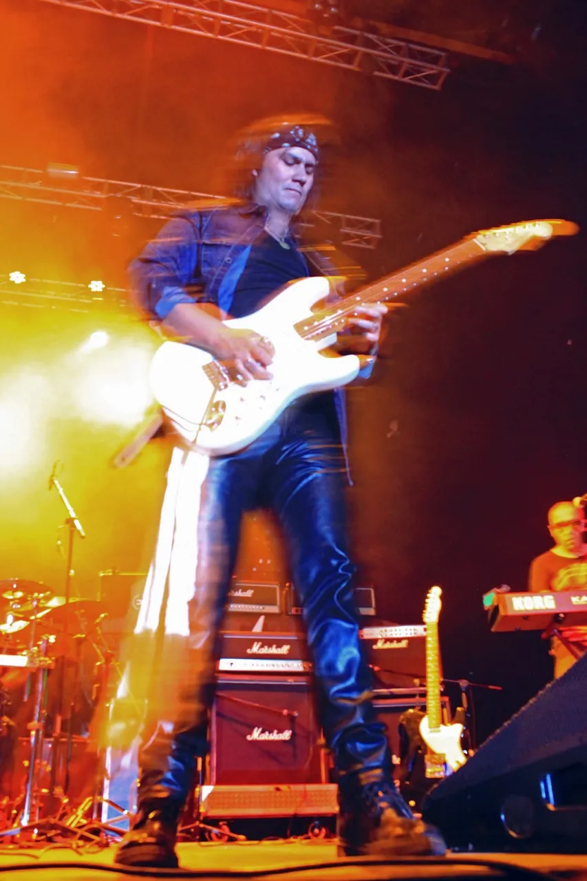 Mateo Acevedo como guitarrista de rock en pleno concierto, iluminado por luces naranjas sobre la tarima.
