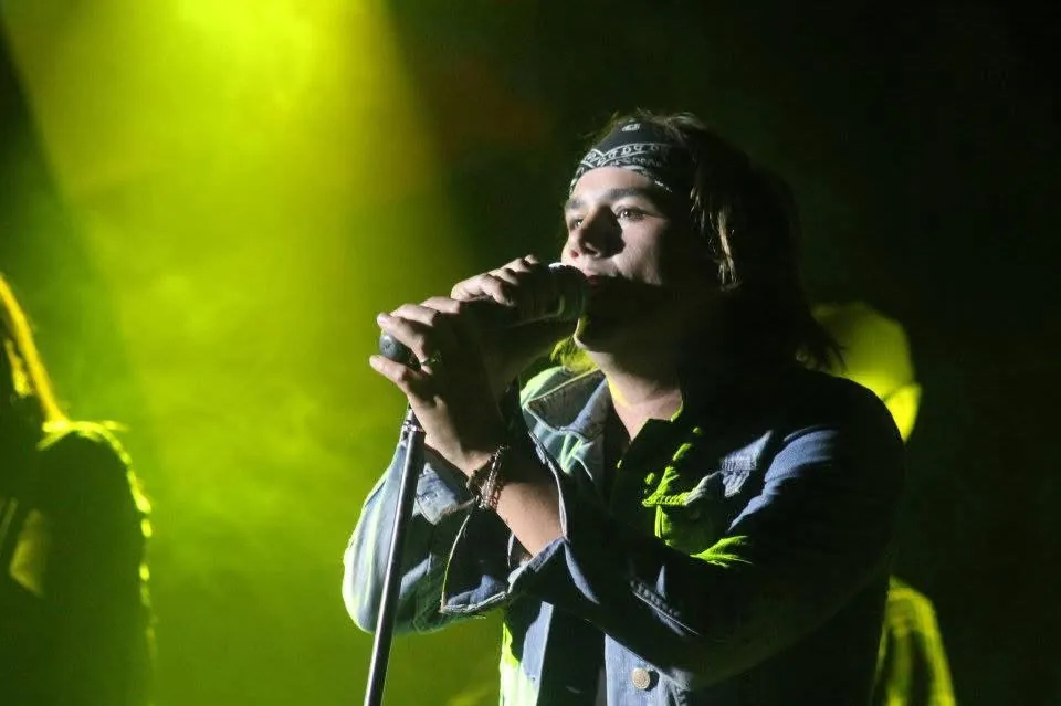 Mateo Acevedo cantando con chaqueta de jean bajo intensas luces verdes durante una presentación en vivo.