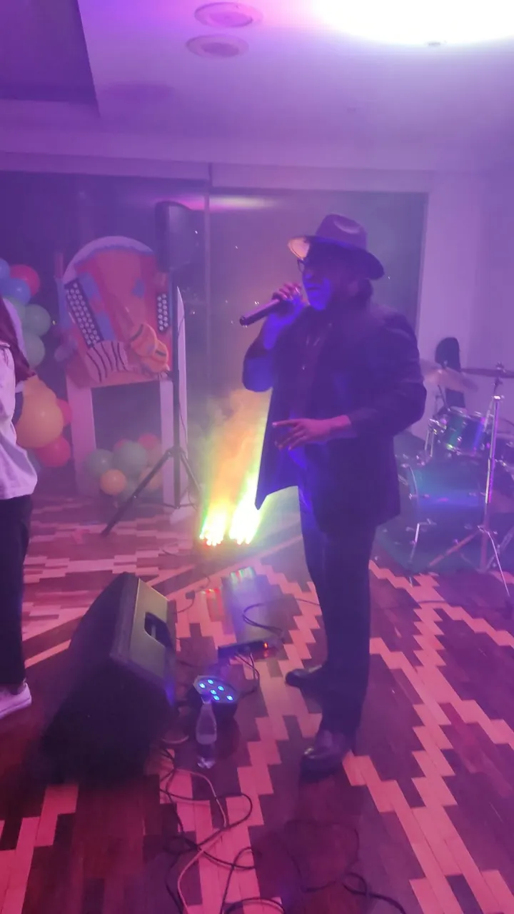Vocalista con sombrero anima un evento en vivo cantando al micrófono entre el humo del escenario.